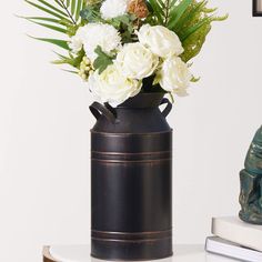 a black vase with white flowers and greenery in it sitting on a table next to a book