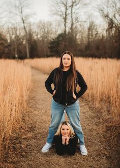 two women are standing in the middle of a field with tall grass and trees behind them