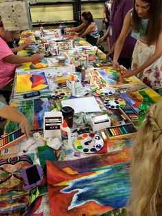 several people are painting on canvass at a long table with other people around them