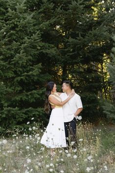 two people standing in the middle of a field with pine trees behind them and white flowers on the ground