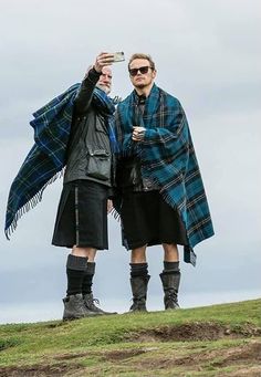 two men in kilts taking a selfie with their cell phone on top of a hill