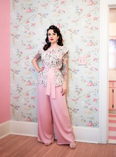 a woman standing in front of a wall wearing pink pants and a white top with flowers on it