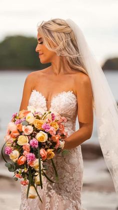 a woman in a wedding dress holding a bouquet and looking at the camera while standing next to water