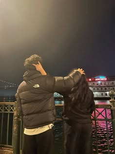 two people standing next to each other in front of a body of water at night