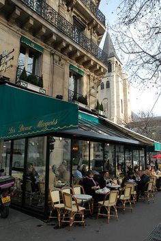 people are sitting at tables in front of a restaurant on the side of the street