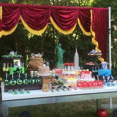 a table topped with lots of cakes and desserts next to a red drapes