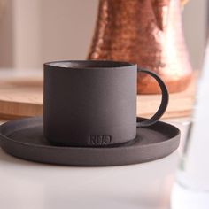 a black coffee cup sitting on top of a saucer next to a copper vase