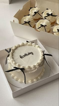 a white cake in a box with black ribbon around it and some cupcakes behind it