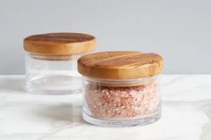 two glass jars with wooden lids on a marble countertop, one containing pink sea salt and the other containing sugar