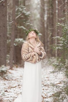 a woman in a white dress and fur coat standing in the snow with her arms crossed