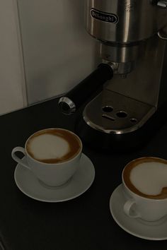 two cups of coffee sitting on top of a black counter next to a silver espresso machine