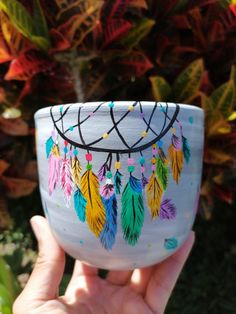 a hand holding a cup with colorful feathers painted on the outside and inside, in front of some plants