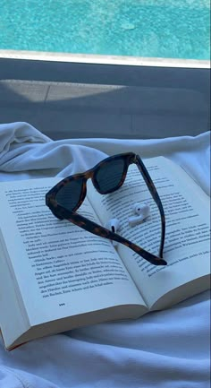 an open book with sunglasses and earbuds laying on it next to a swimming pool