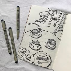 an open notebook with some drawings on it and two pens in front of it next to each other