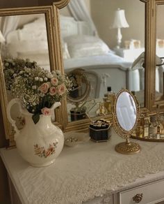 a white dresser topped with a mirror and vase filled with flowers next to a gold framed mirror