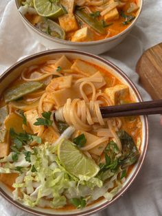 two bowls filled with noodles and vegetables on top of a white table cloth next to a wooden spoon