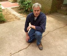 an older man squatting down on the ground in front of a house with his dog