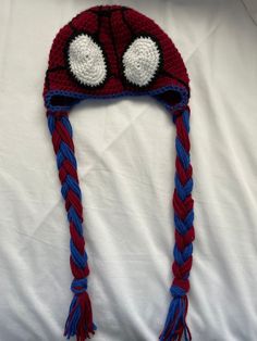 a crocheted spiderman hat with eyes and braids on the front, sitting on a white sheet