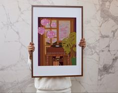 a person holding up a framed painting with a window and table in front of them