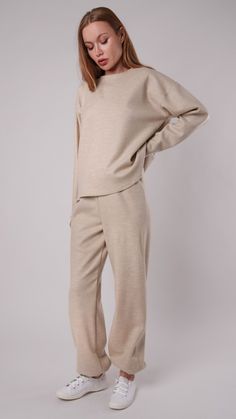 a woman standing in front of a white background wearing a beige sweater and sweatpants