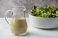 a glass pitcher filled with dressing next to a bowl of lettuce and a salad