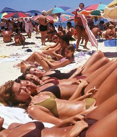 many people are laying on the beach in bikinis and sunbathers with umbrellas