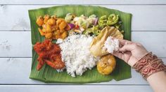 a person holding a plate with rice, meat and veggies on it in front of a banana leaf