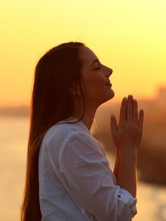 a woman with her hands together in front of the sun at sunset, praying or meditating