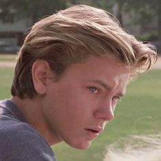 a young man with blonde hair and blue eyes stares into the distance while standing in front of a grassy area