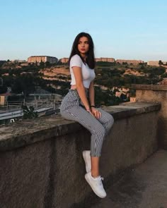 a woman sitting on top of a stone wall next to a cityscape with buildings in the background