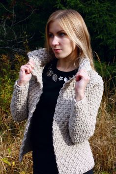 a woman in black shirt and white jacket standing next to grass with trees behind her