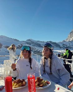 two women sitting at a table with food and drinks in front of snow covered mountains