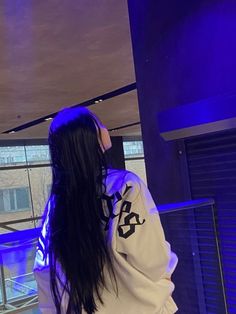 a woman with long black hair standing in front of a blue lit window looking at something