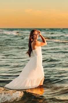 a woman standing in the ocean wearing a white dress and holding her hand to her head