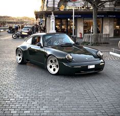 a black sports car parked in front of a building on a brick road with people walking by