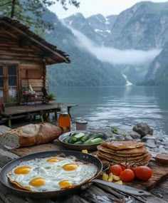 some food is sitting on a wooden table by the water with mountains in the background