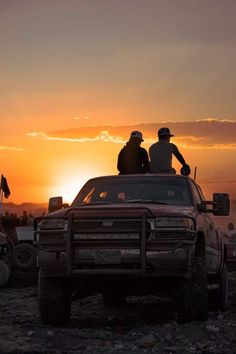 two men sitting on the back of a pickup truck in front of an orange sunset