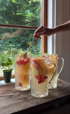 two glasses filled with liquid and fruit on top of a wooden table next to a window