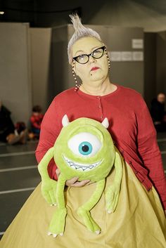 a woman in a red shirt is holding a green stuffed animal with big blue eyes