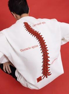 a man sitting on the ground with his back turned to the camera, wearing a white shirt with red embroidery