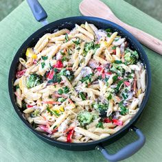 a skillet filled with pasta and vegetables on top of a green cloth next to a wooden spoon