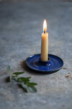 a single candle is lit on a blue plate with green leaves around it and resting on the ground