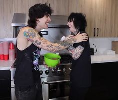 two people standing in a kitchen with tattoos on their arms and hands, one holding a green bowl