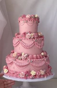 a three tiered cake with pink frosting and flowers on it, being held by someone's hand