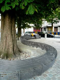 a curved metal bench sitting next to a tree on a city street with cars parked in the background