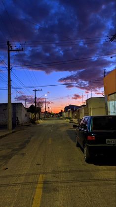 a black car parked on the side of a road at dusk with clouds in the sky