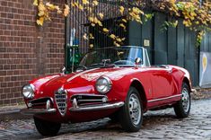 an old red sports car parked in front of a brick building on a cobblestone street