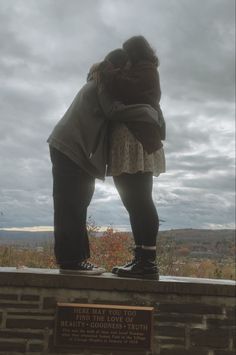 two people hugging each other on top of a stone wall in front of a cloudy sky