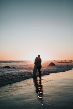 a man and woman standing on top of a beach next to the ocean at sunset