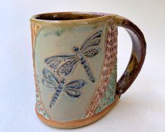 a ceramic mug with dragonflies on it's side, sitting on a white surface